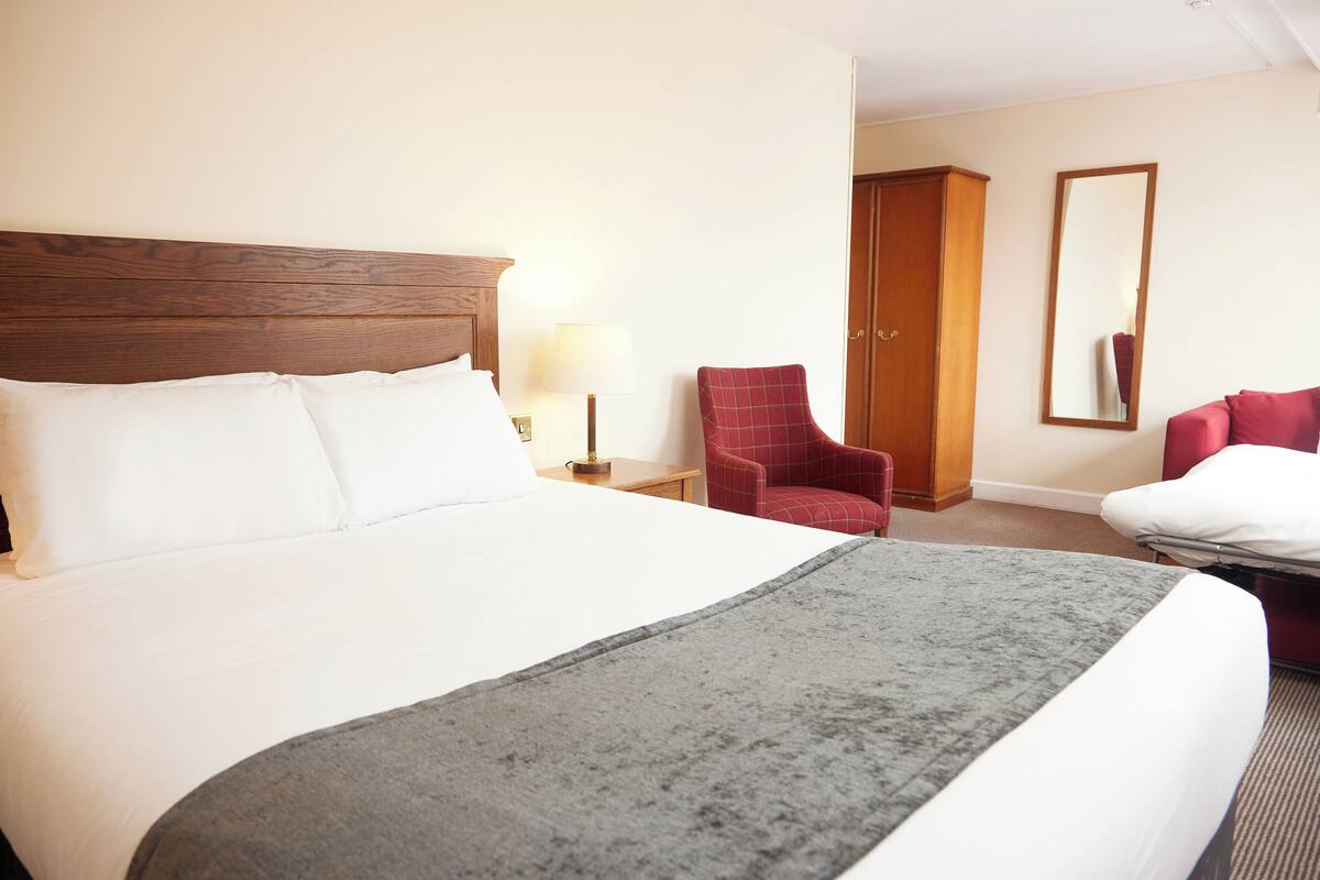 A hotel room with a large bed, a wooden headboard, a red armchair, a desk with a lamp, and a wardrobe.