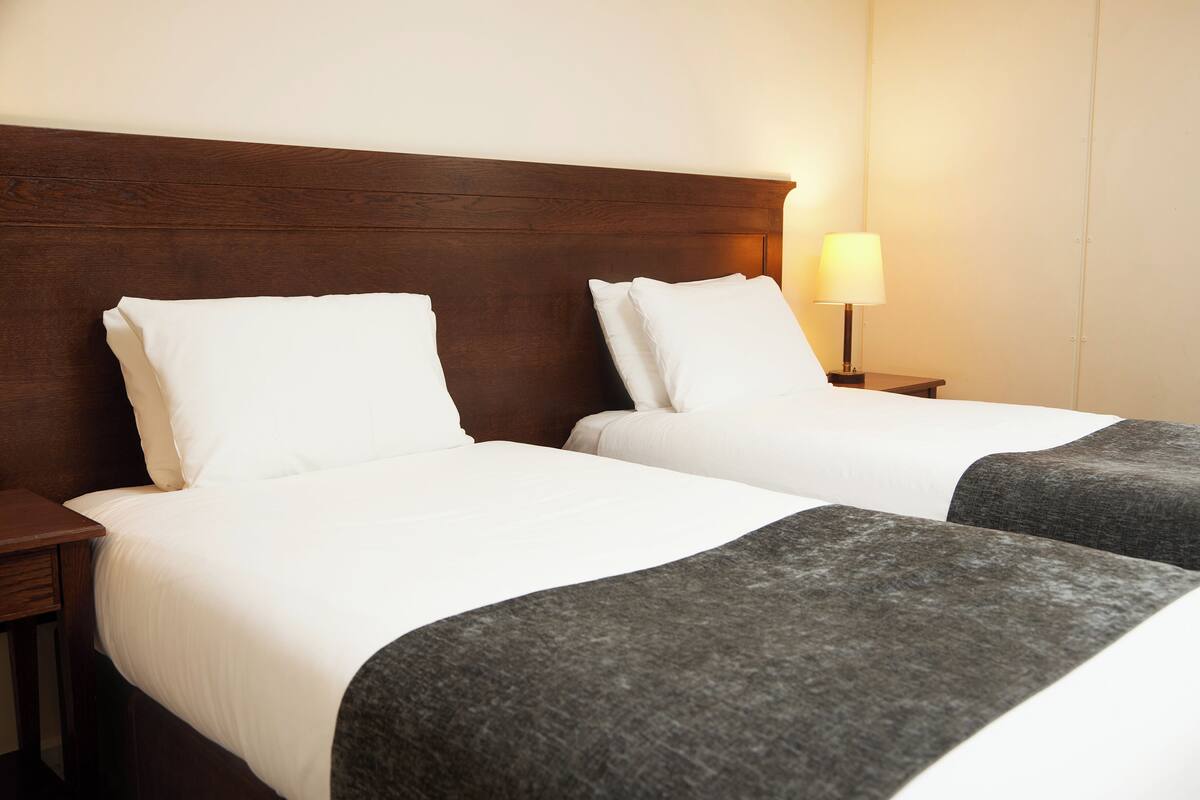 A hotel room with two beds, wooden headboard, white bedding, and a bedside lamp.
