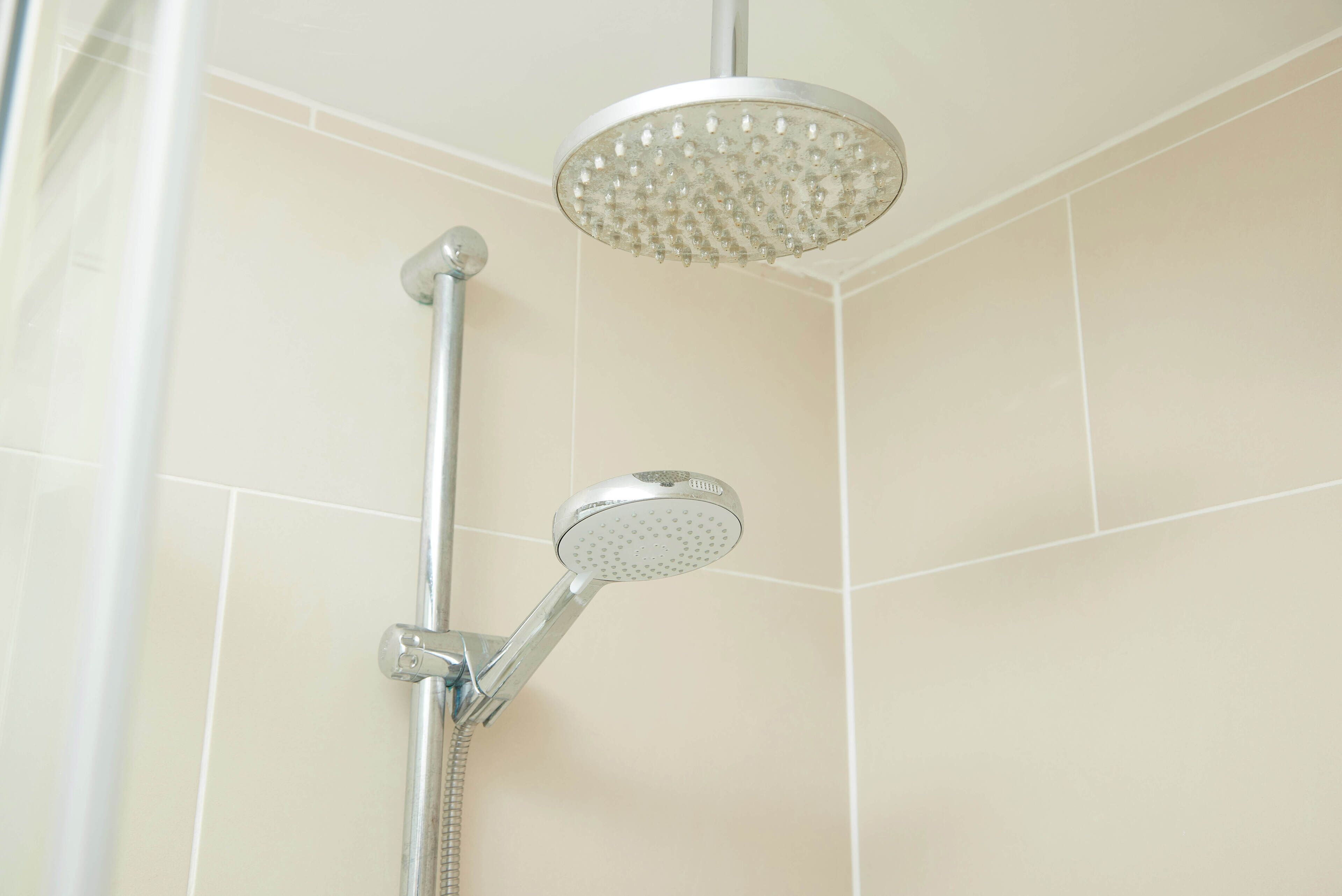 A showerhead with a handheld attachment, mounted on a wall with beige tiles.