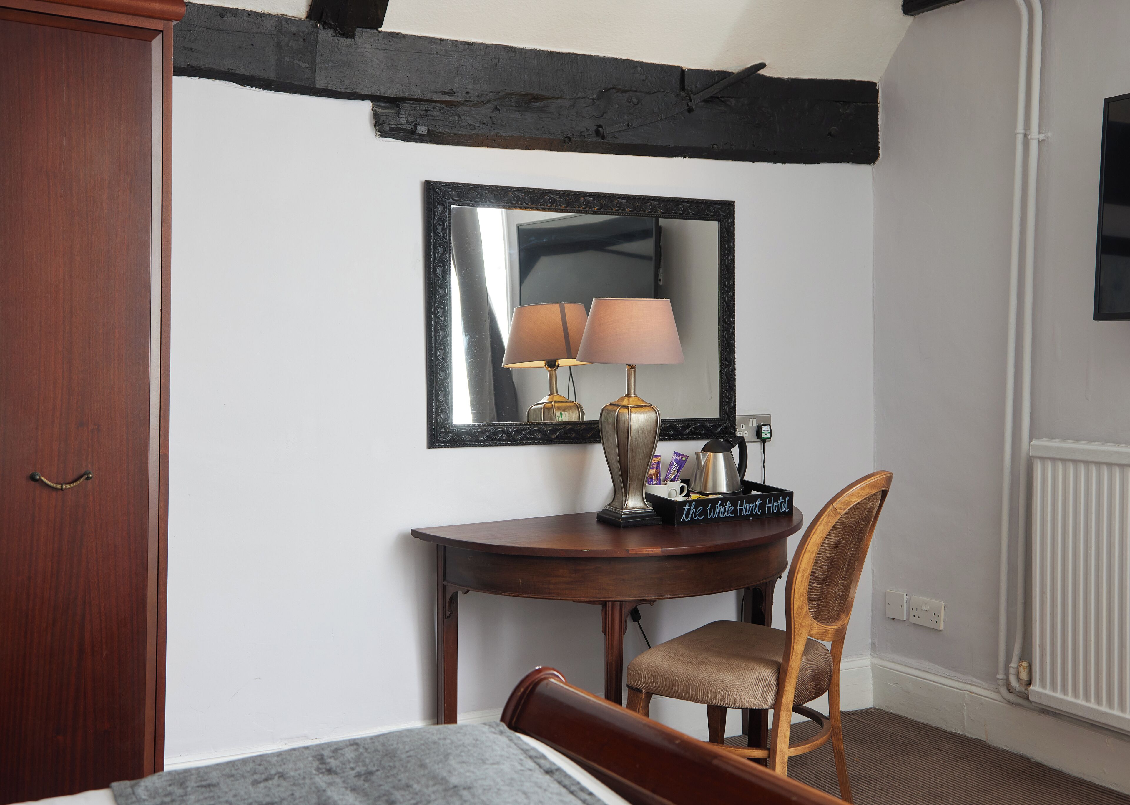 Een hotelkamer met een houten bureau, een stoel, een bed, een spiegel, een lamp en een dienblad met een koffiezetapparaat en een mok.