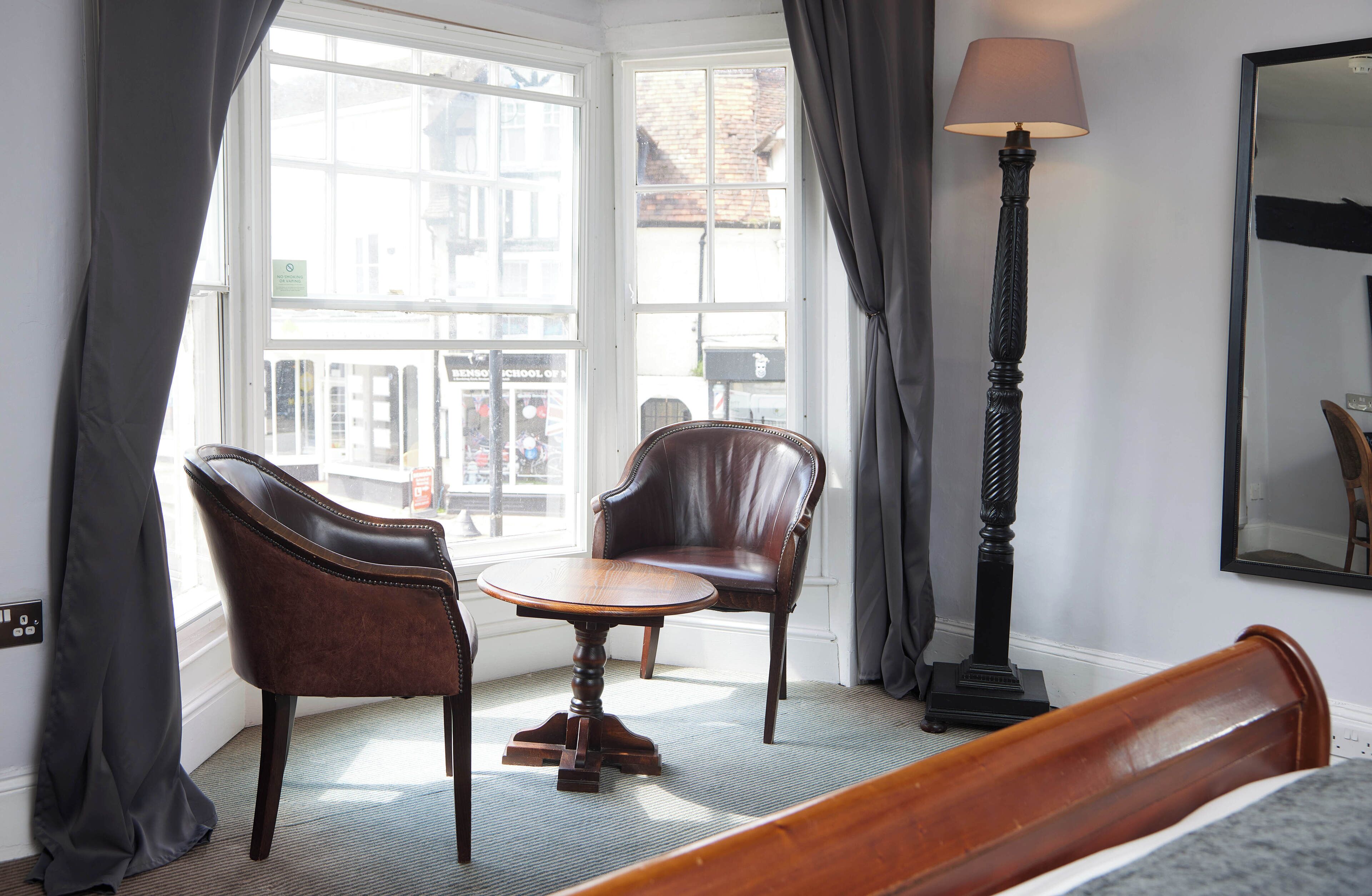 A hotel room with a large window, two leather chairs, a round wooden table, a floor lamp, and a bed.