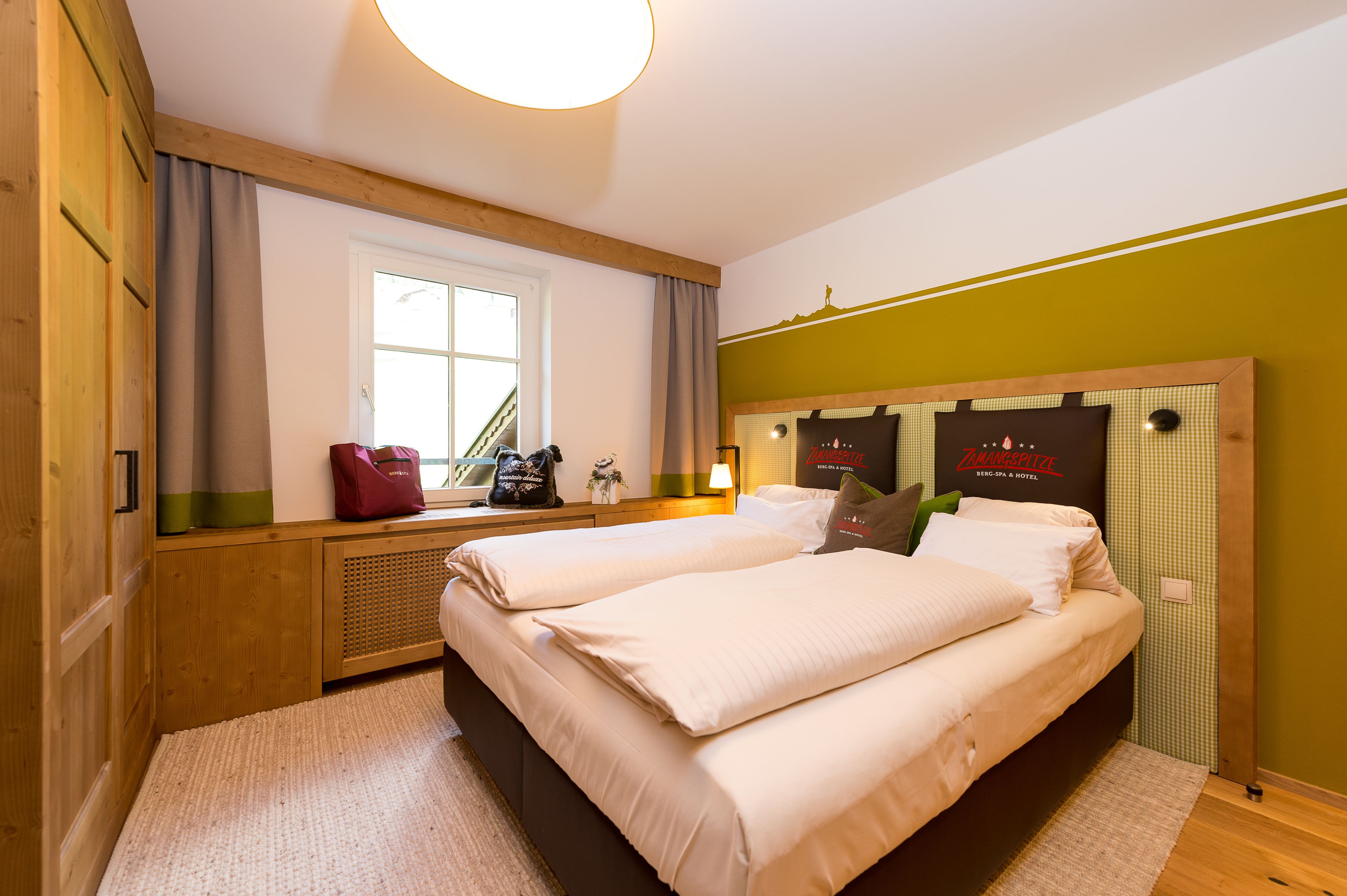 Une chambre d’hôtel avec deux lits, une fenêtre avec des rideaux, une tête de lit en bois et une tête de lit fixée au mur portant le nom de l’hôtel.