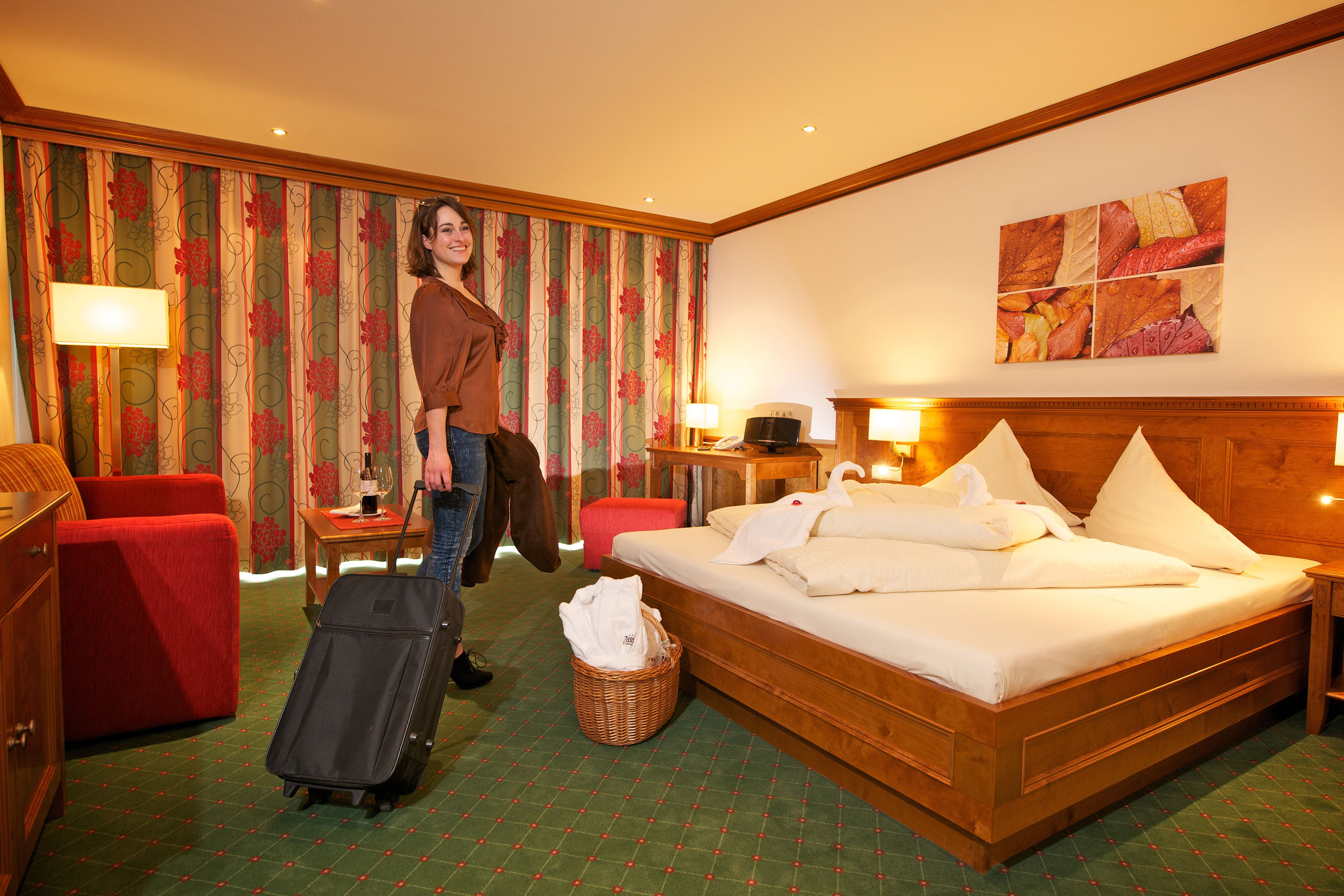 A hotel room with a bed, a desk, a chair, a suitcase, and a wall with a floral pattern.