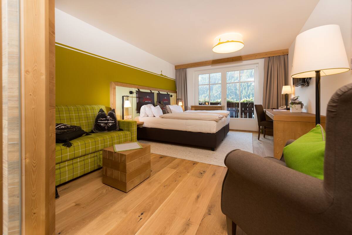 Een hotelkamer met een bed, een groene bank, een houten bureau en uitzicht op het omliggende landschap.