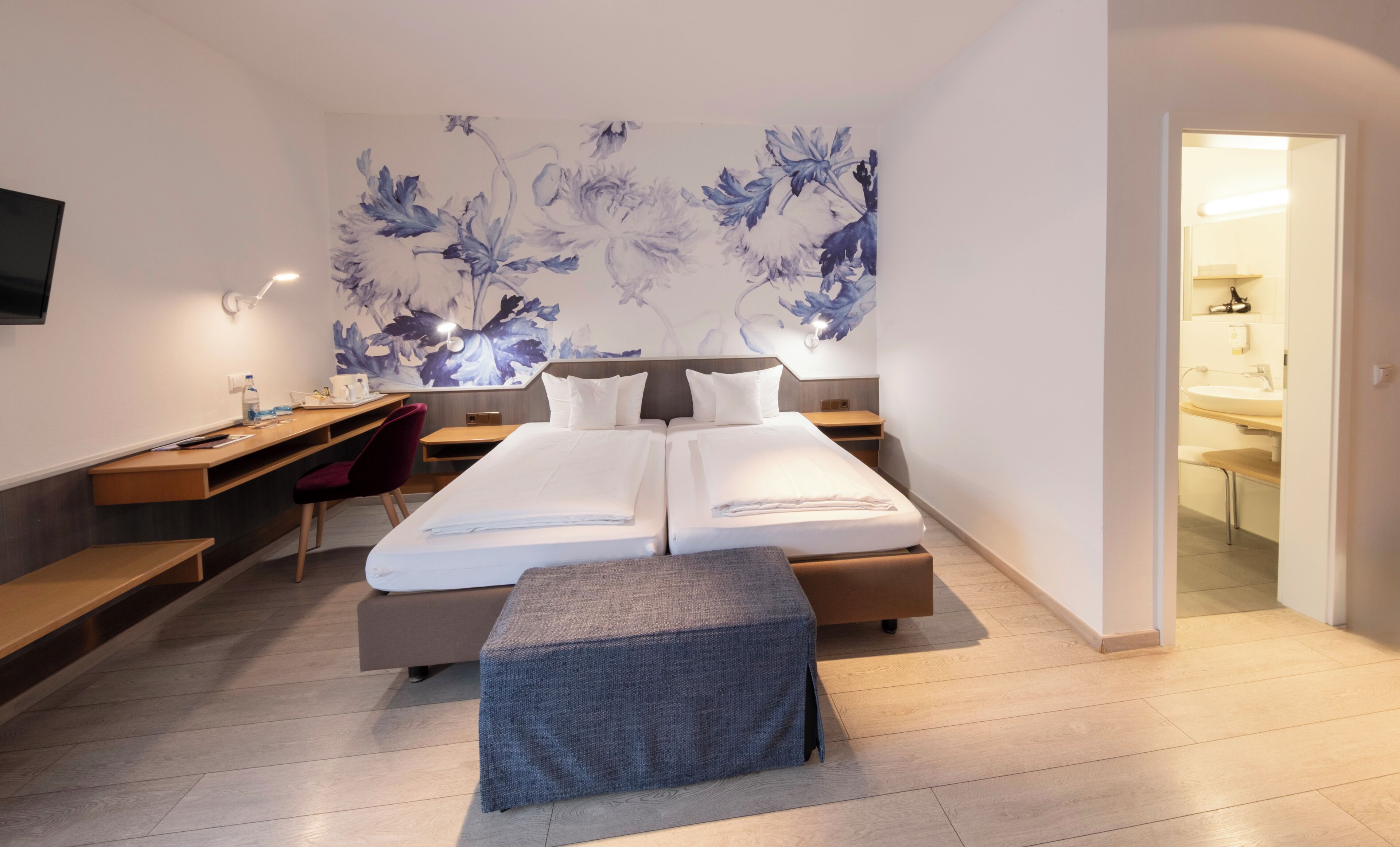 A modern hotel room with a bed, desk, chair, and a large floral artwork on the wall.