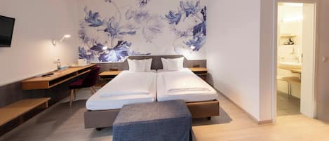 A modern hotel room with a bed, desk, chair, and a large floral artwork on the wall.