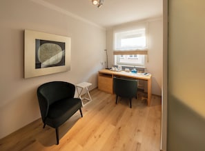Ein Zimmer mit Schreibtisch, zwei Stühlen, einem Heizkörper, einem Fenster mit Jalousien und einem gerahmten Kunstwerk an der Wand.