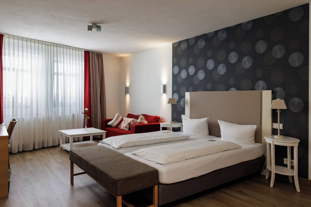 A hotel room with two beds, a red sofa, a small table, and a lamp.