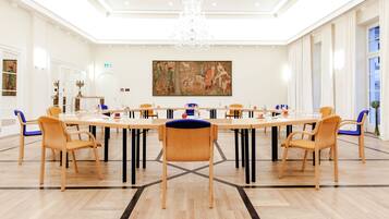 A conference room with a long table, wooden chairs, a chandelier, and a large painting on the wall.