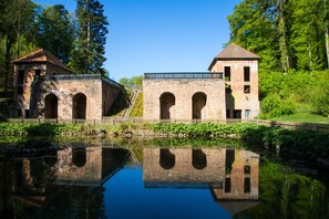 Un edificio histórico de piedra con ventanas de arco y una torre central, rodeado de abundante vegetación y una masa de agua tranquila que refleja su estructura.