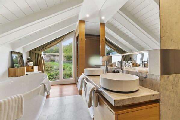 Un baño moderno con una bañera independiente, dos lavamanos redondos, un espejo grande y vistas a zonas verdes a través de las ventanas.