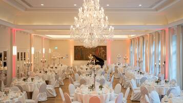 Une salle de banquet avec des tables rondes, des chaises blanches, des lustres et un piano.