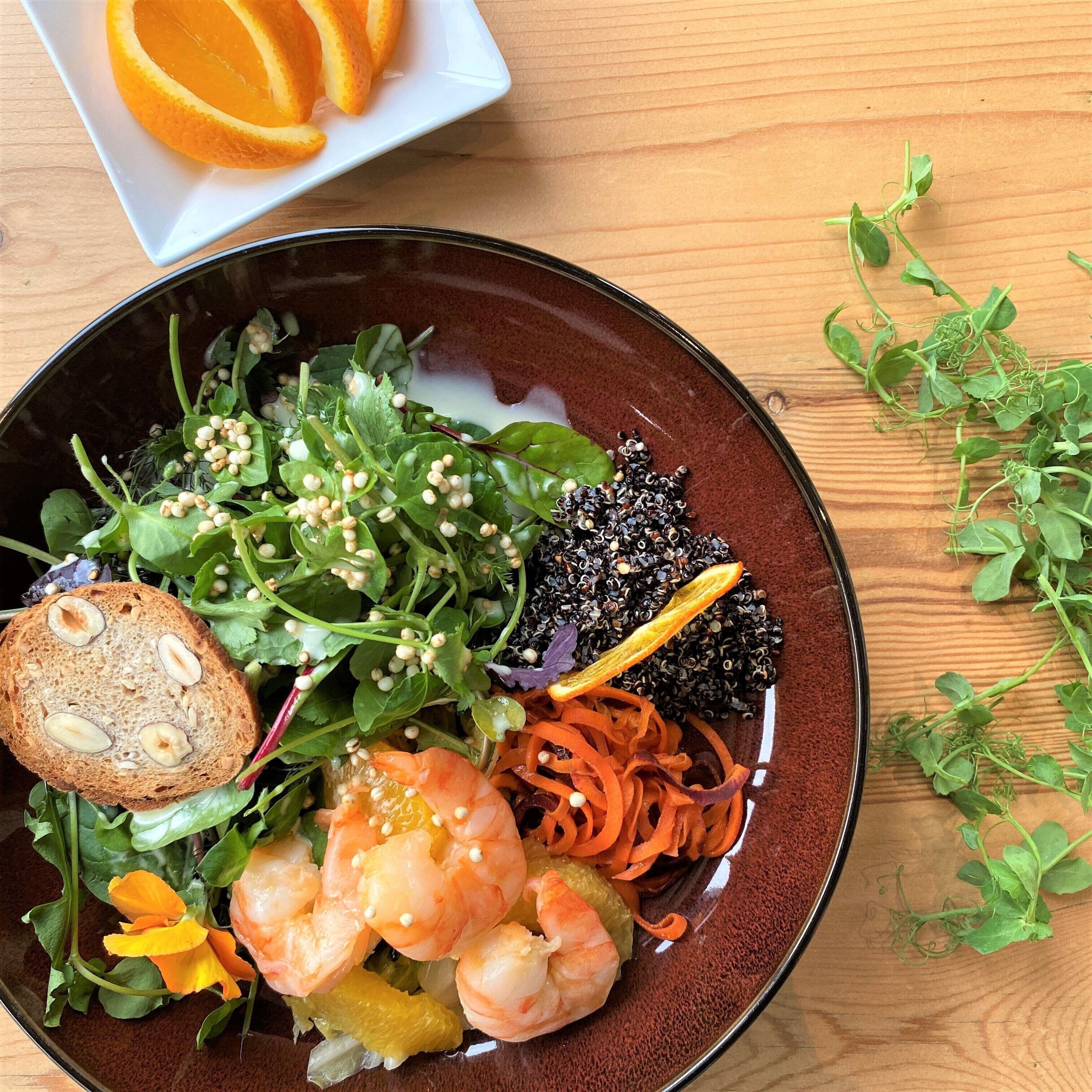 A bowl of mixed salad with various vegetables and shrimp, accompanied by a slice of bread and a plate of orange slices.