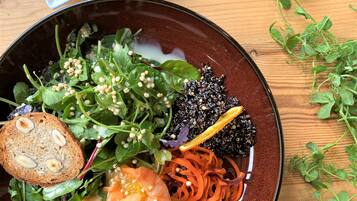 Eine Schüssel gemischter Salat mit verschiedenen Gemüsesorten und Garnelen, dazu eine Scheibe Brot und eine Portion Orangenscheiben.