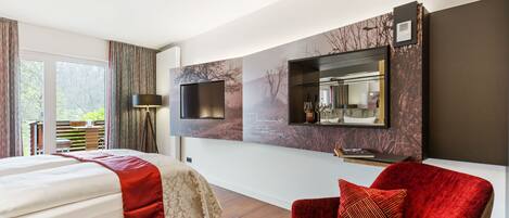Una habitación de hotel moderna con una cama grande, un sillón rojo, un televisor montado y un ventanal con cortinas.