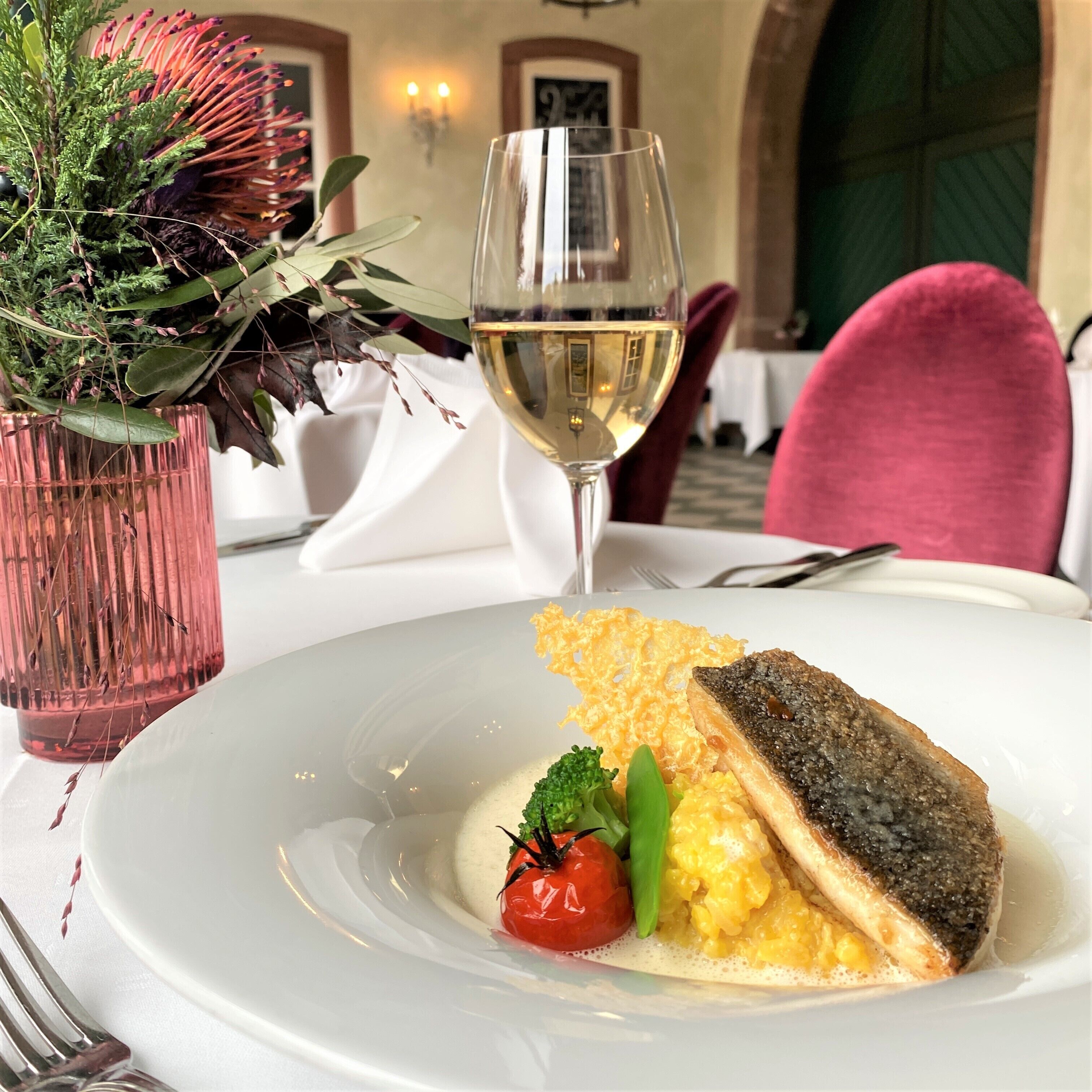Una mesa de comedor con un plato blanco de comida, una copa de vino blanco y un florero rosa con plantas verdes.