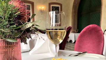 Una mesa de comedor con un plato blanco de comida, una copa de vino blanco y un florero rosa con plantas verdes.