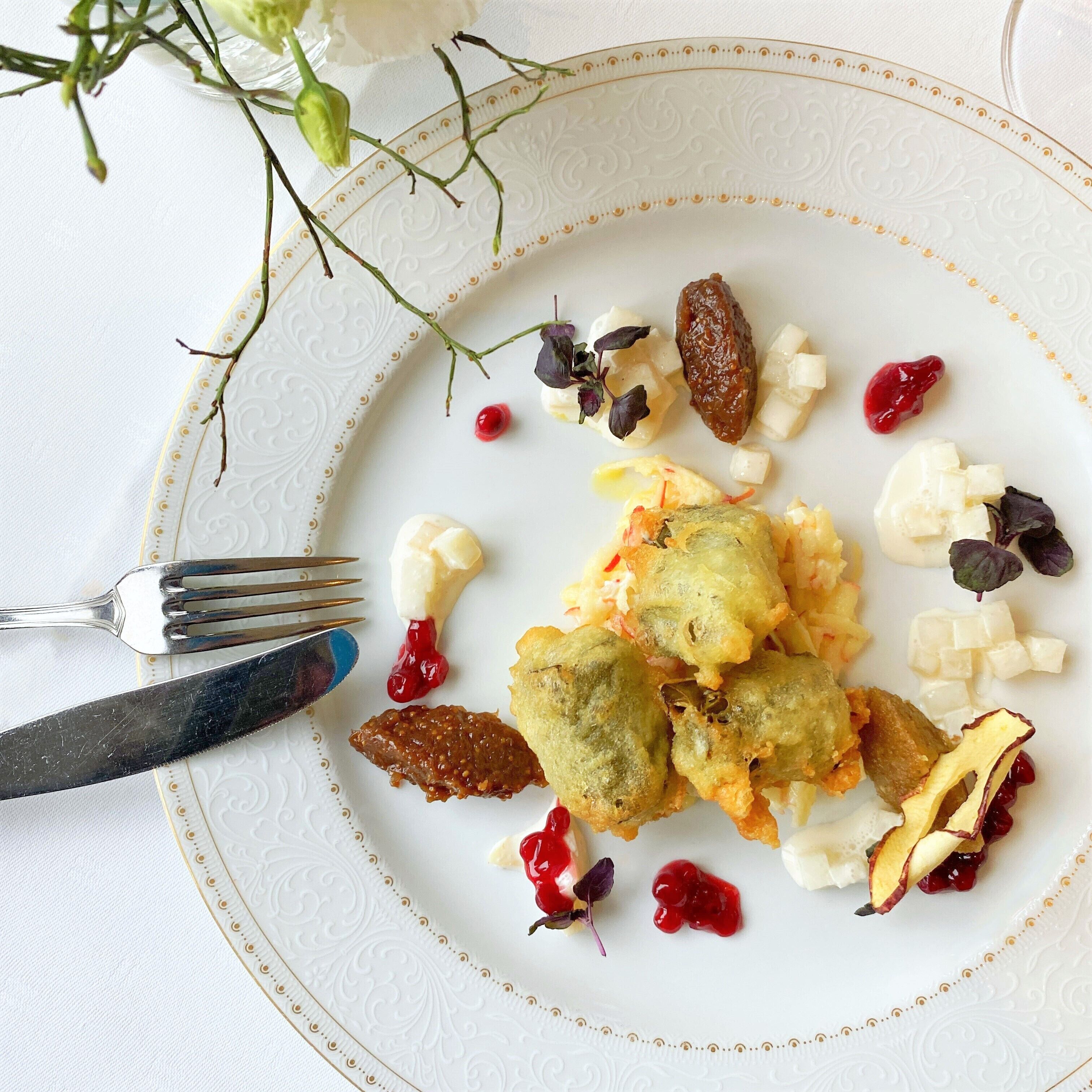 Een bord eten met diverse ingrediënten, waaronder gefrituurde stukken, kaas en gedroogd fruit, met een vork en mes op een wit bord met een gouden rand.