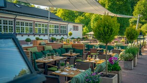 An outdoor cafe with wooden tables and green cushioned seating, surrounded by potted plants and a white building in the background. - Romantik Hotel Landschloss Fasanerie (Zweibruecken)