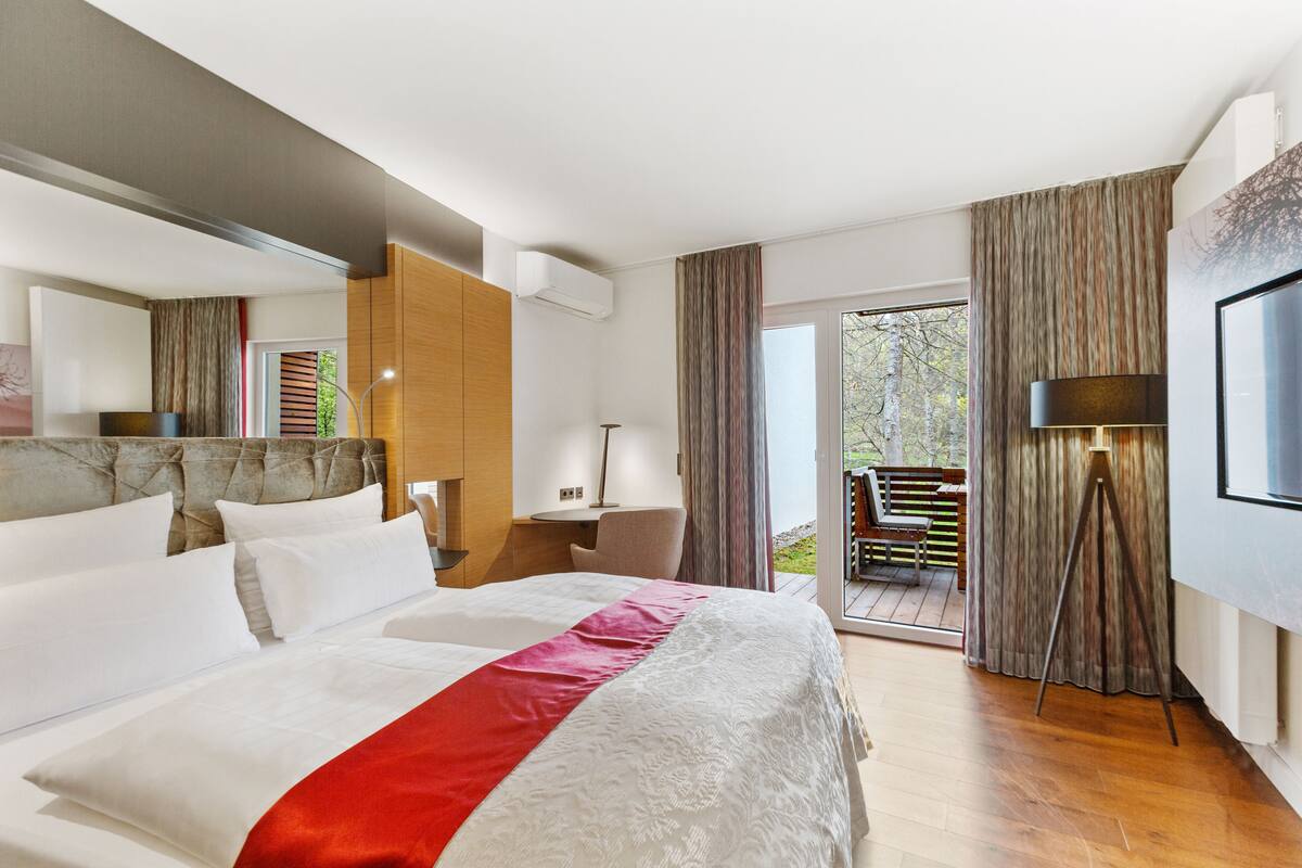 A modern hotel room with a large bed, a desk, and a balcony with outdoor seating.
