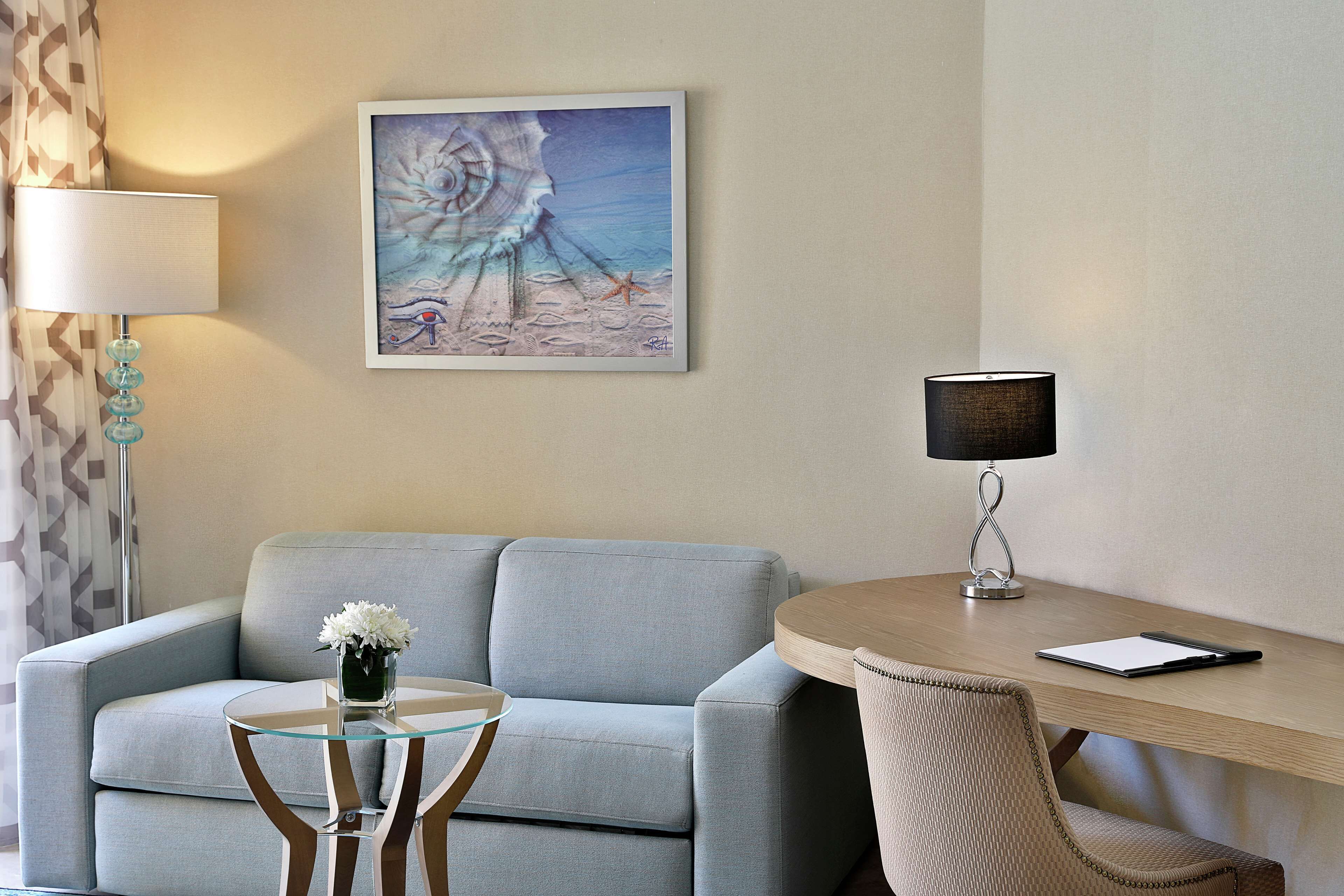 A modern hotel room with a light blue sofa, a glass coffee table with a vase of flowers, a wooden desk with a lamp, and a framed artwork on the wall.