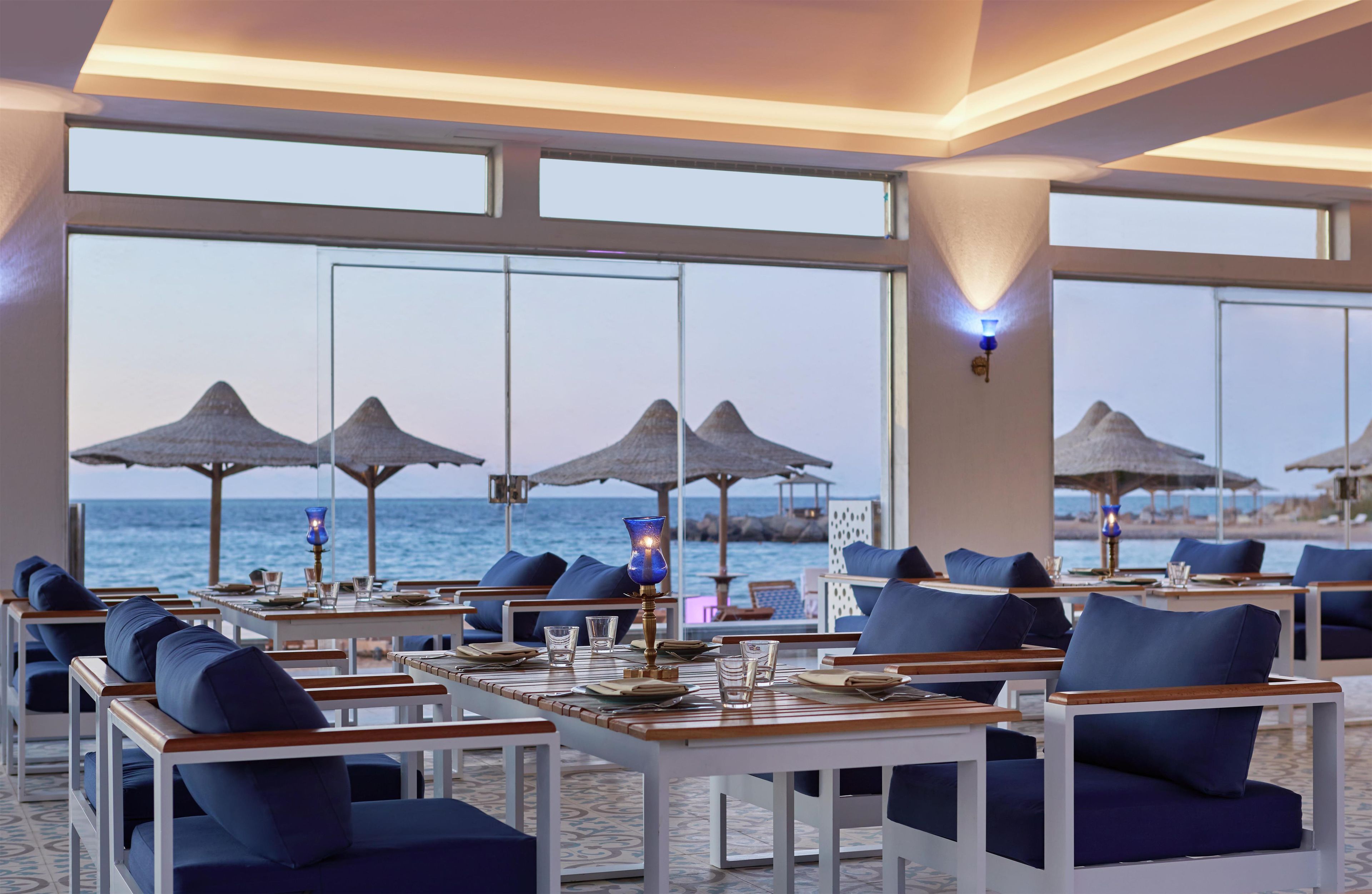 A seaside restaurant with tables and chairs, large windows, and umbrellas.