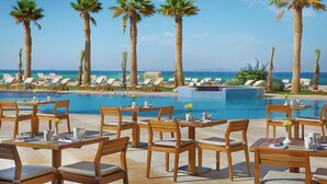 A poolside dining area with wooden tables and chairs, a clear view of the ocean, and palm trees. - Hilton Hurghada Plaza (Hurghada)