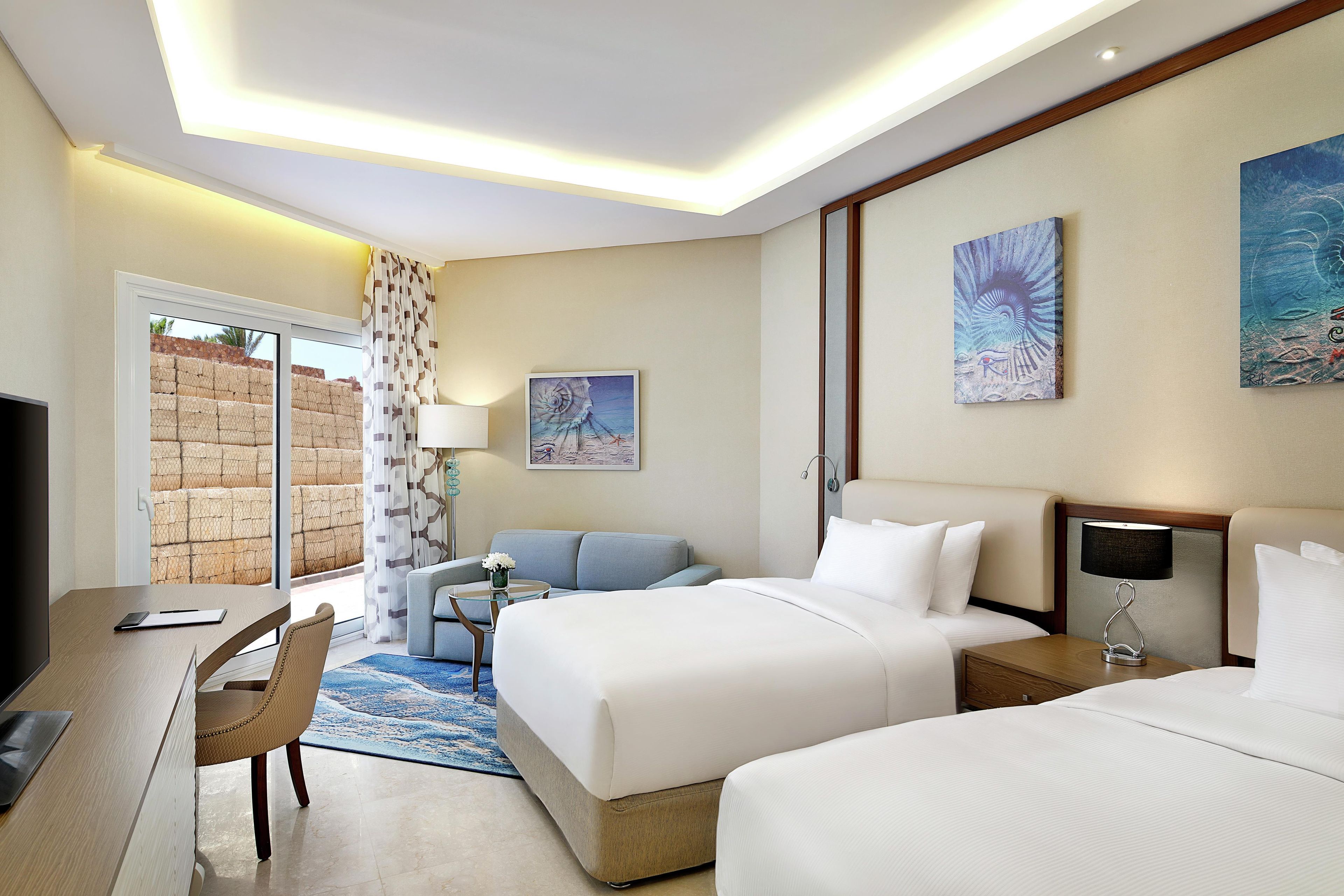 A hotel room with a bed, bedside tables, a desk, a chair, a television, and artwork on the walls.