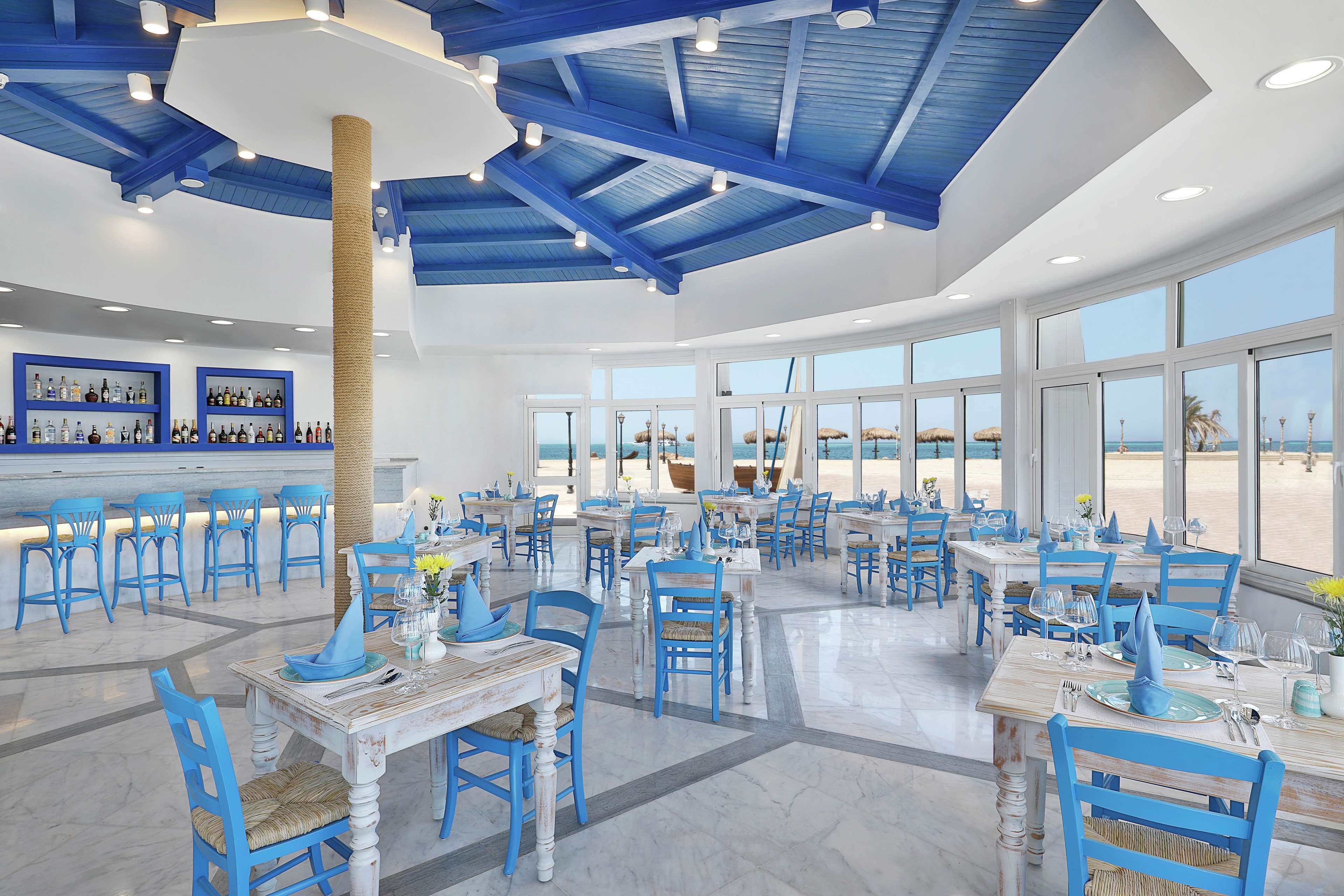 A spacious dining area with blue chairs and tables, large windows offering a view of the beach, and a bar area with shelves of bottles.