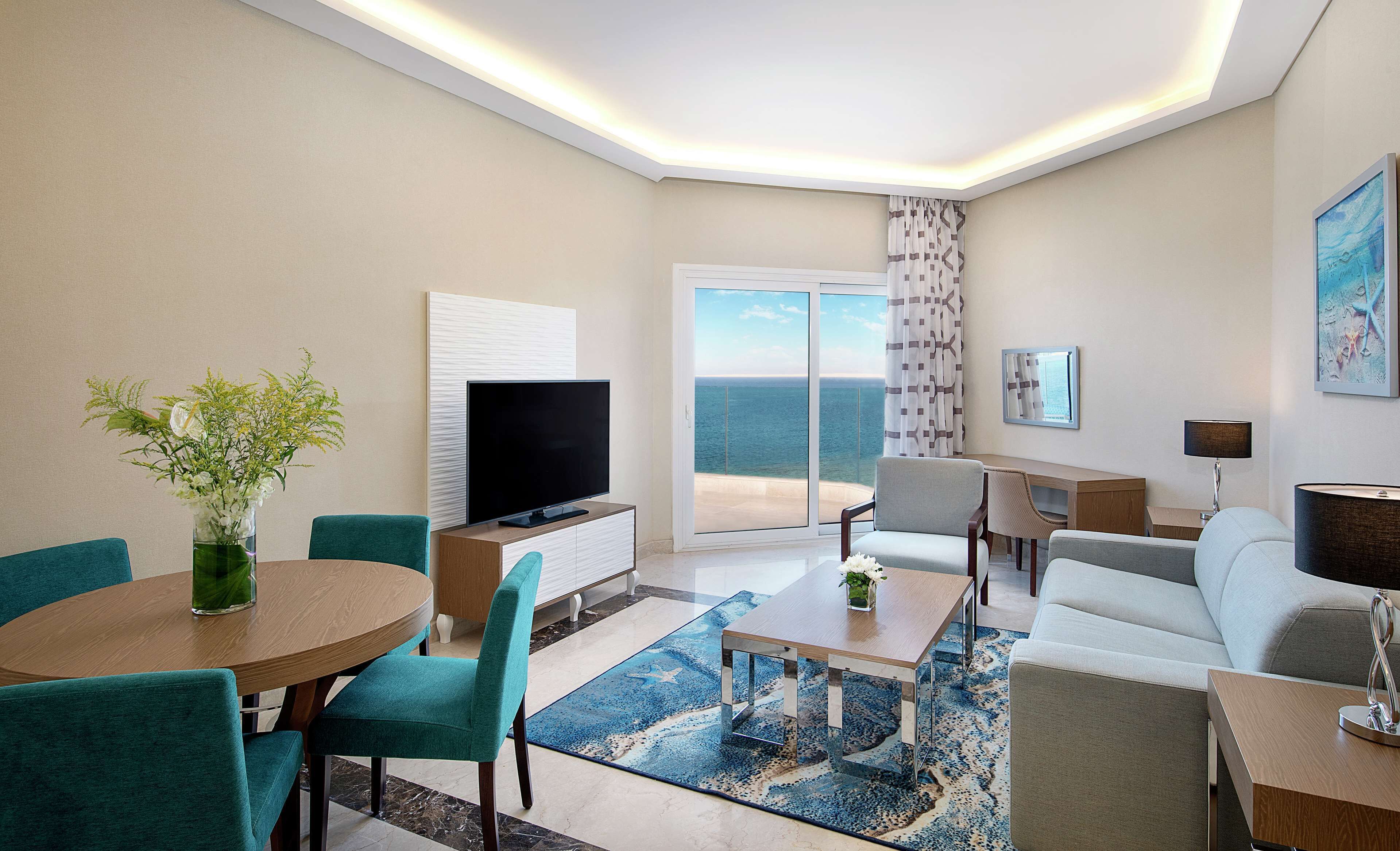 A modern hotel room with a balcony, a flat-screen TV, teal chairs, a wooden coffee table, and a large window offering a view of the ocean.