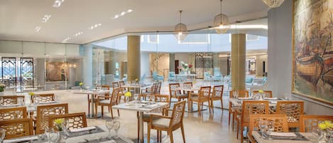 A spacious dining area with multiple tables and chairs, a high ceiling with recessed lighting, and a large mural on the wall.