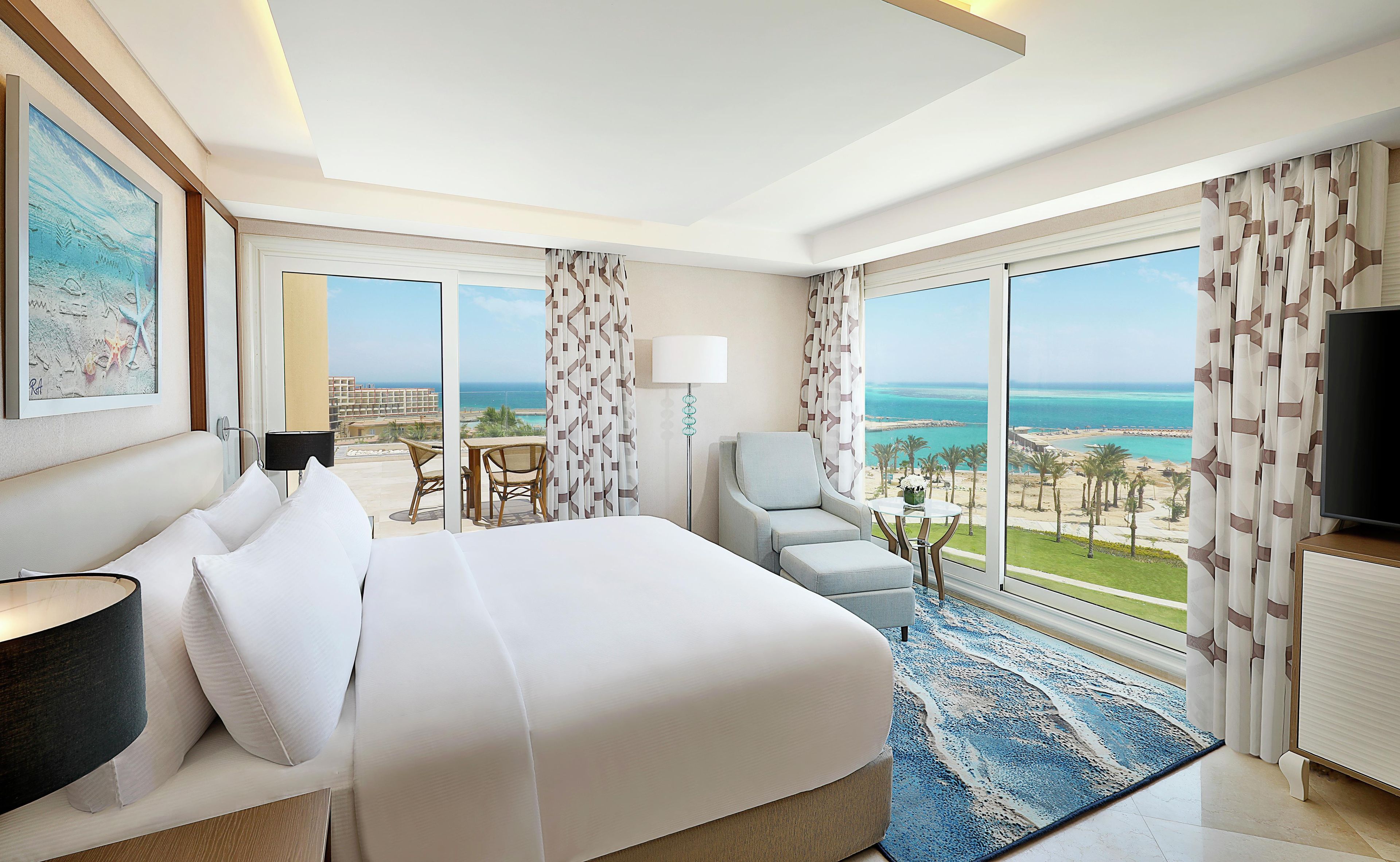 Ein Hotelzimmer mit Bett, Schreibtisch, Sessel, Fernseher und großen Fenstern mit Meerblick.