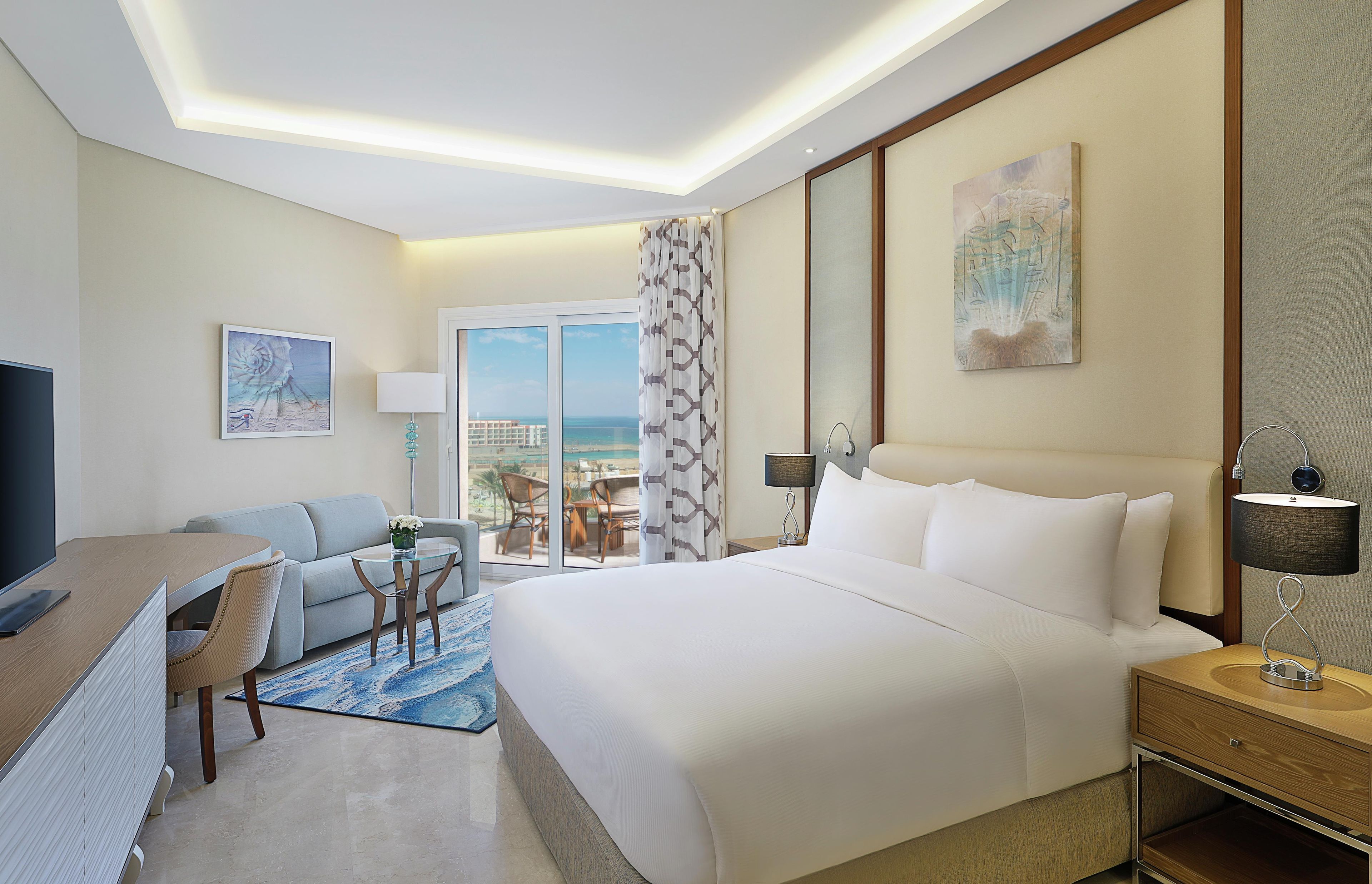A hotel room with a large bed, a desk with a computer, a chair, a TV, and a view of the beach.