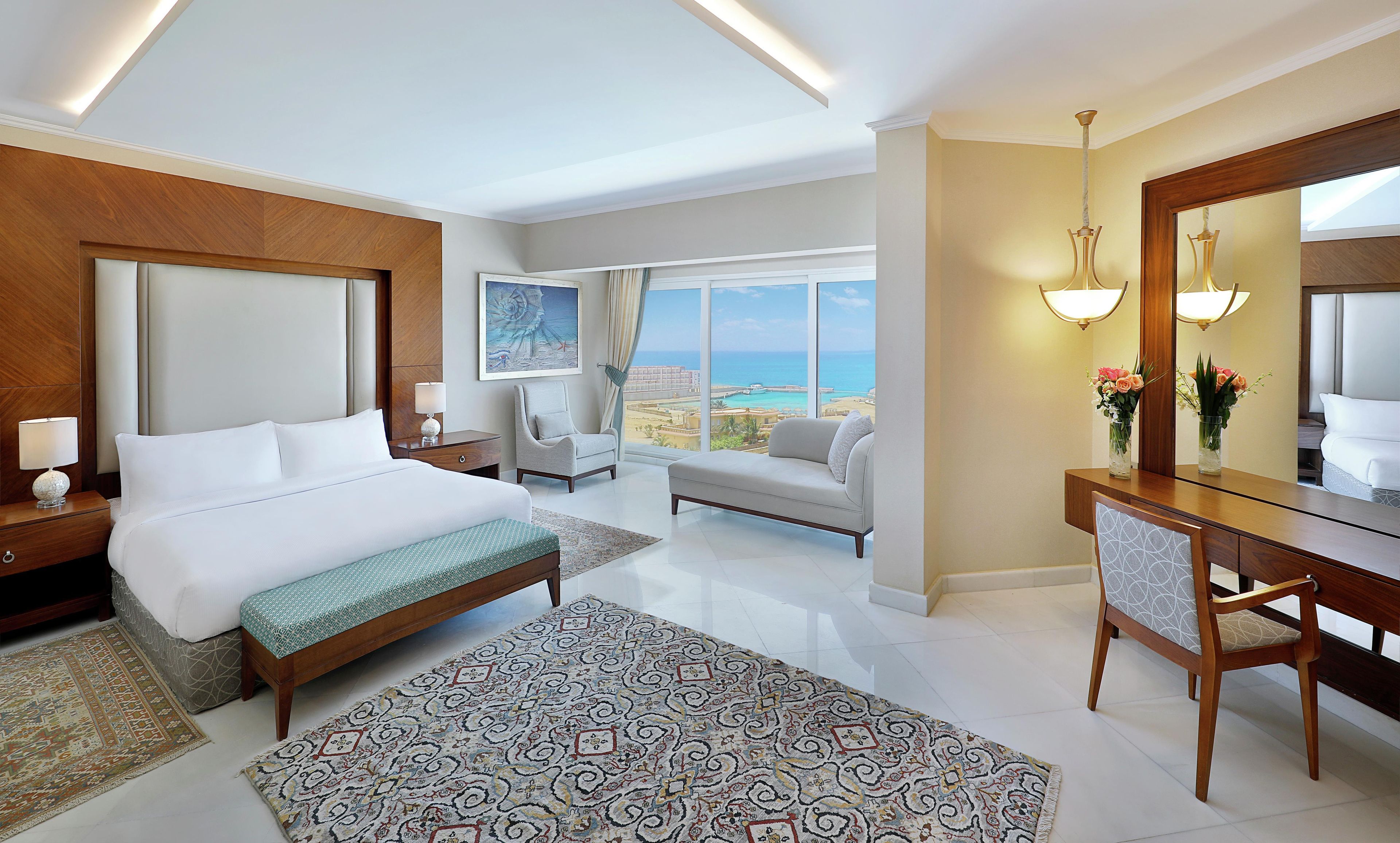 A hotel room with a bed, a sitting area, a desk, and a large window offering a view of the beach.