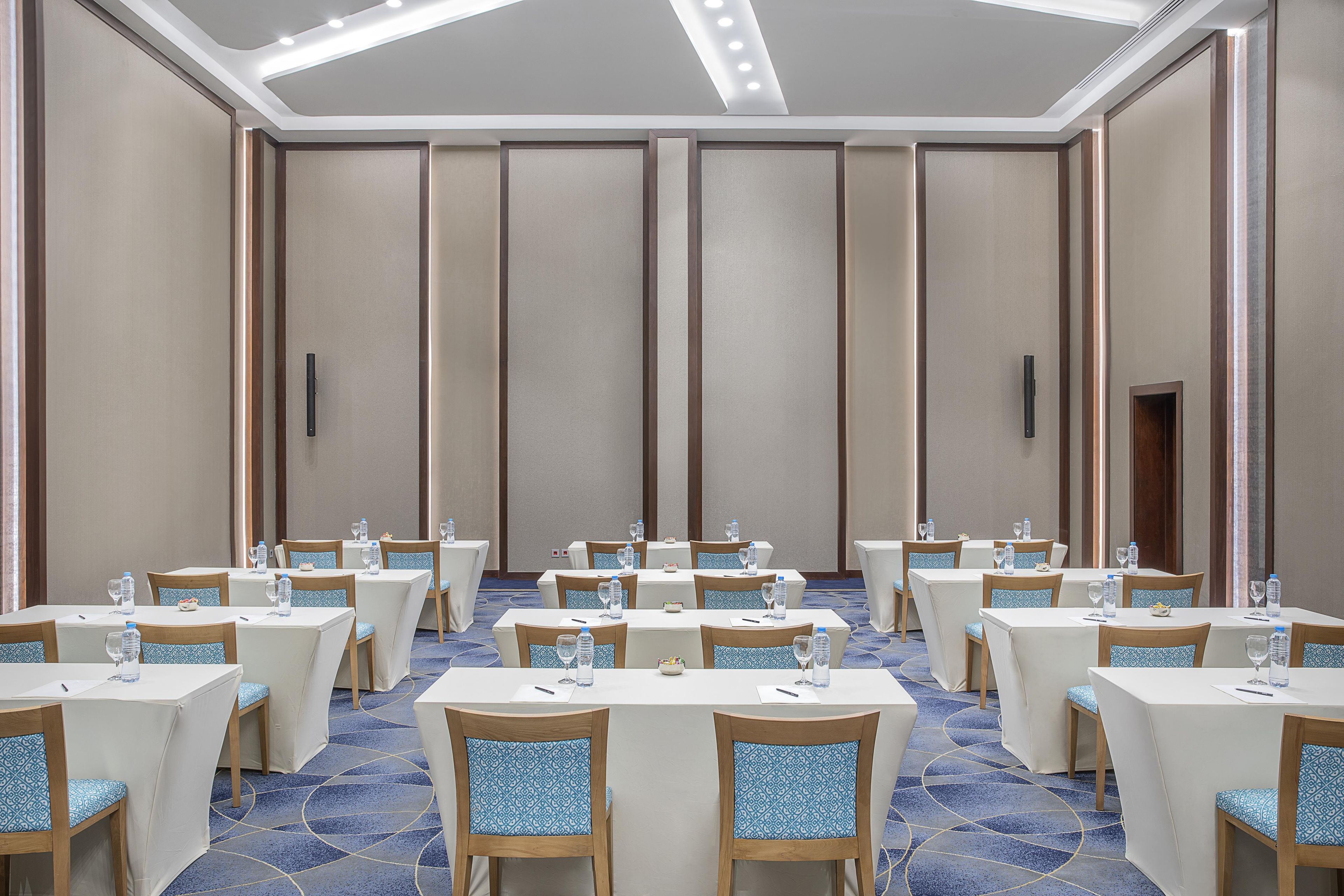Une salle de conférence équipée de tables et de chaises, dotée d’un sol recouvert de moquette et d’un plafond haut avec des luminaires encastrés.
