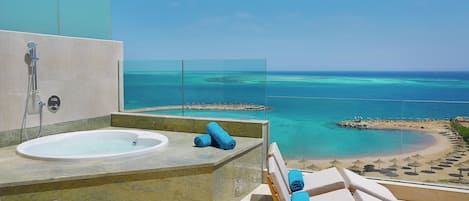A balcony with a view of a beach and ocean, a bathtub, and lounge chairs with towels.