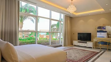 A hotel room with a large bed, a TV, a chandelier, and a view of palm trees and outdoor furniture.