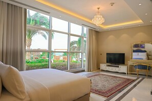 Habitación de hotel con una cama grande, televisor, candelabro y vistas a palmeras y mobiliario exterior.