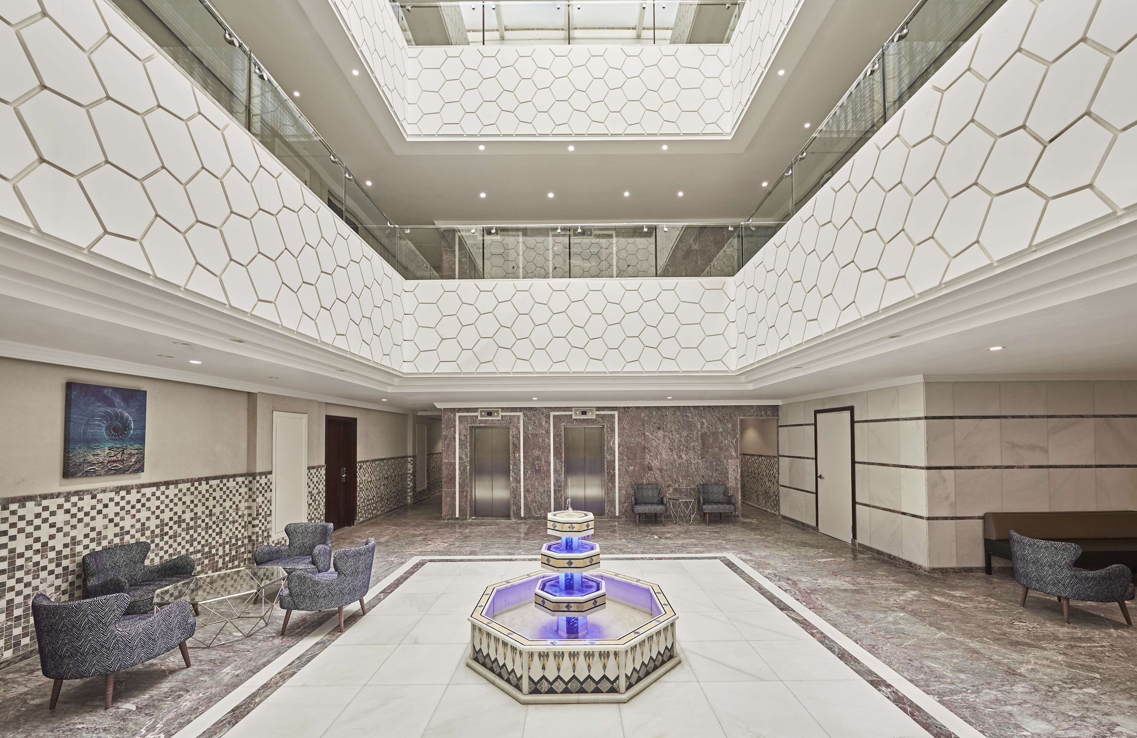 A spacious lobby with a central fountain, seating area, elevators, and a high ceiling with a geometric pattern.