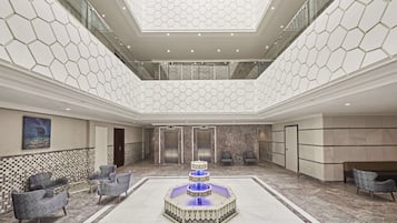 A spacious lobby with a central fountain, seating area, elevators, and a high ceiling with a geometric pattern.