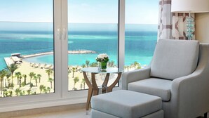 A room with a large window offering a view of a beach and ocean. There is a glass table with a vase of flowers and a lamp. - Hilton Hurghada Plaza (Hurghada)