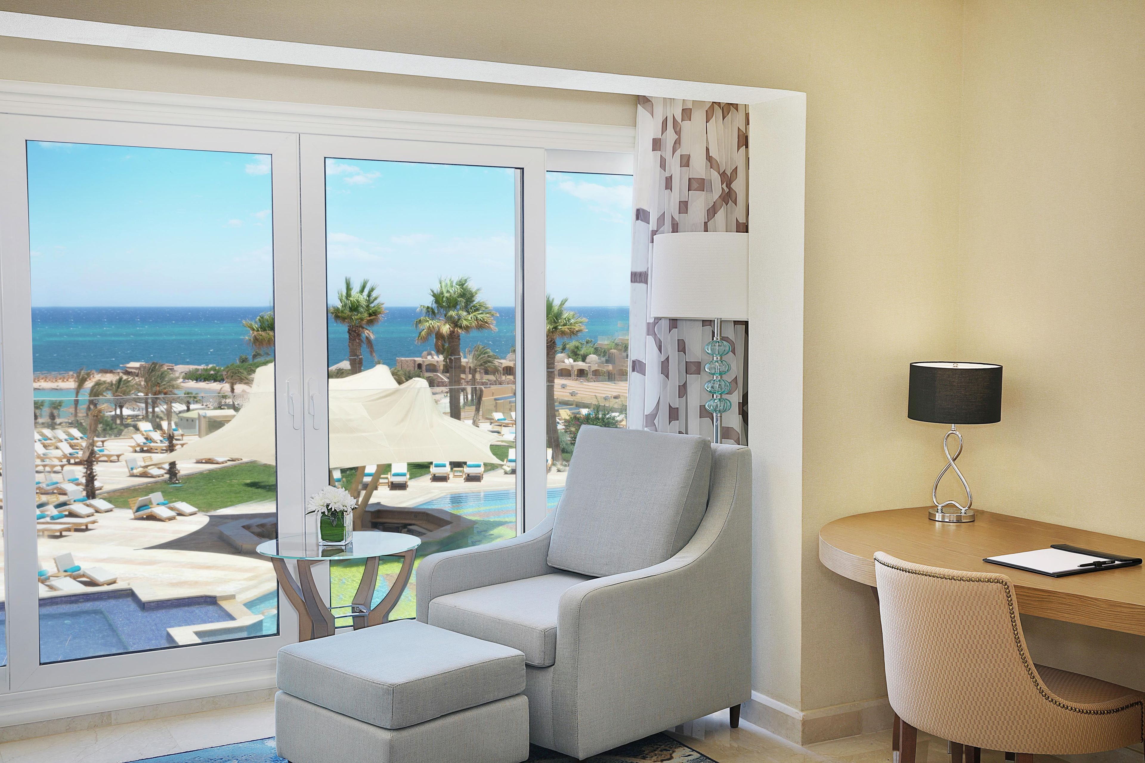 A room with a view of the beach, a chair, a small table, and a lamp.