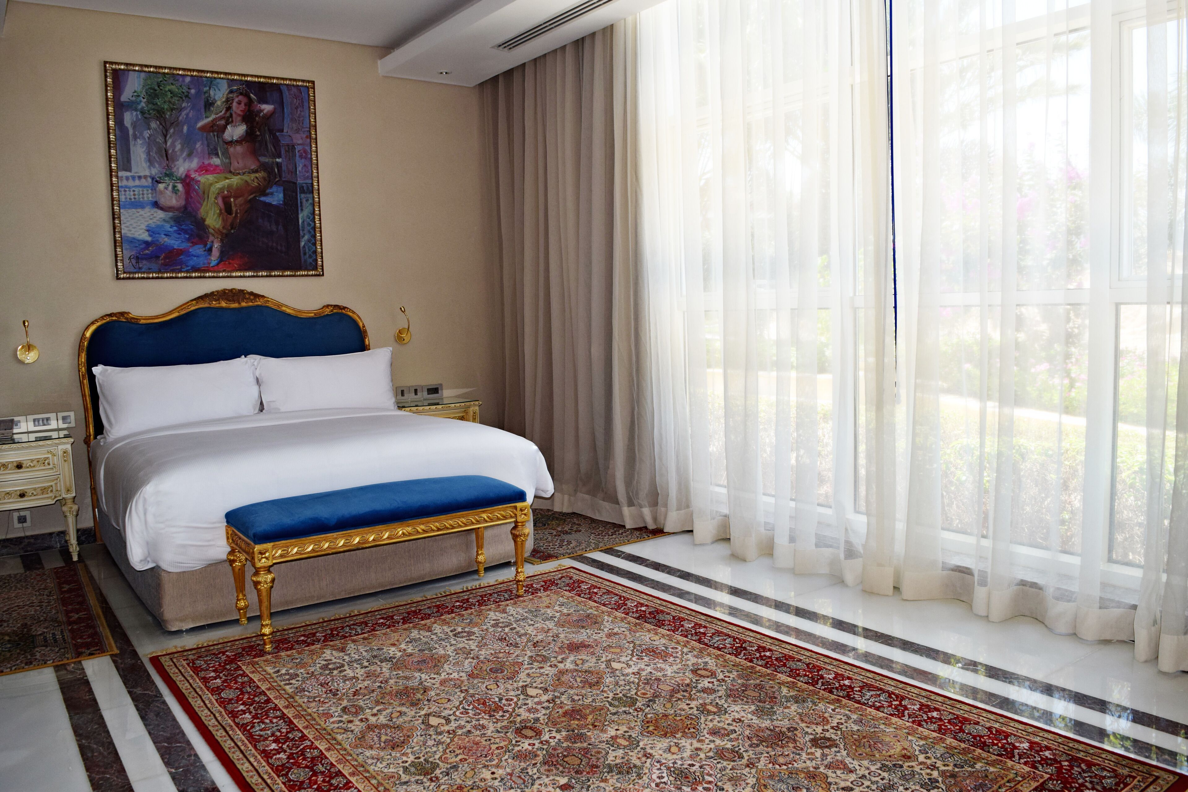 Un dormitorio con una cama grande, un banco azul, un tapete de diseño rojo y beige, un cuadro en la pared y cortinas traslúcidas.