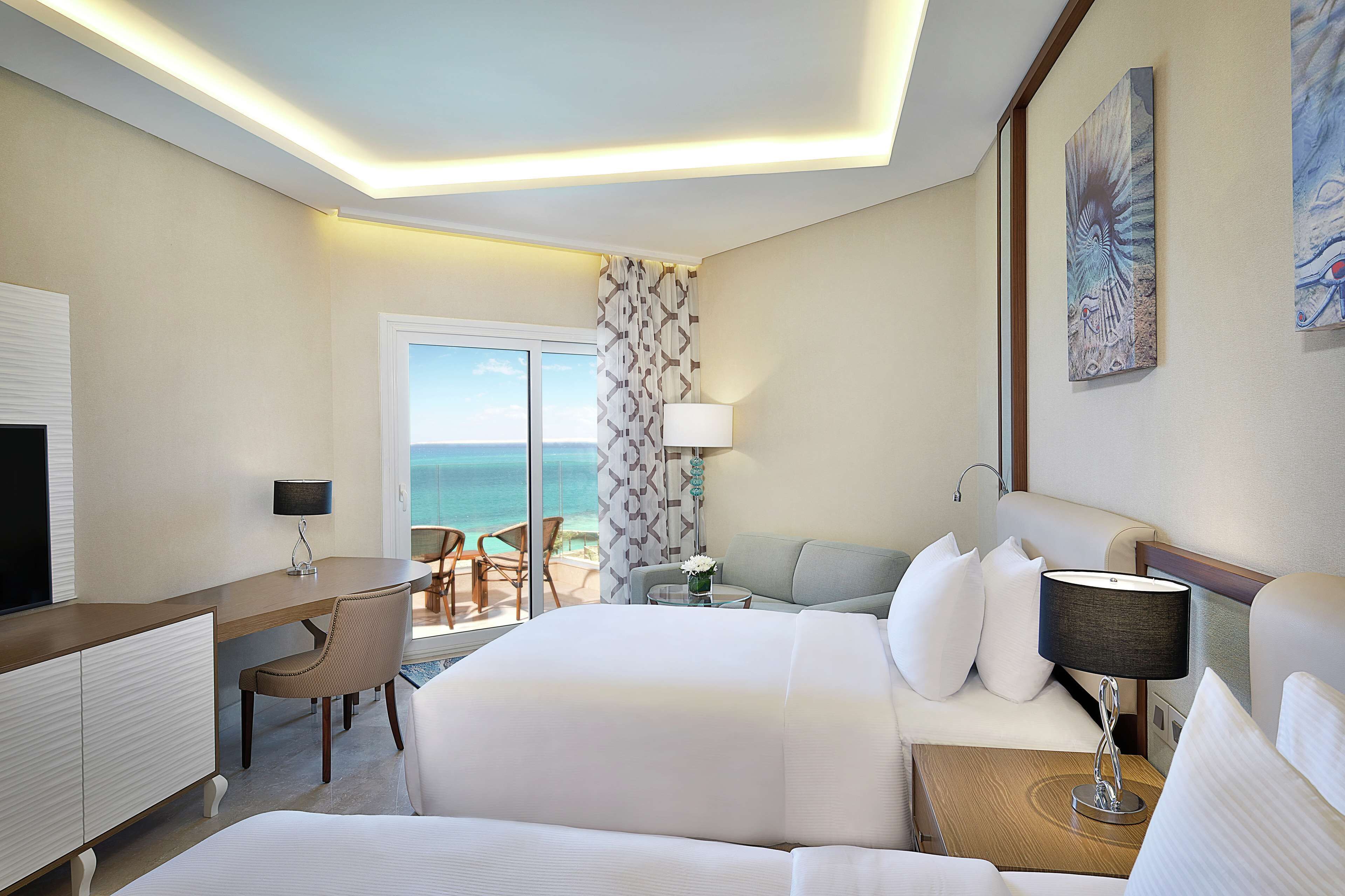 Een hotelkamer met een bed, een bureau, een stoel, een lamp en uitzicht op de oceaan.