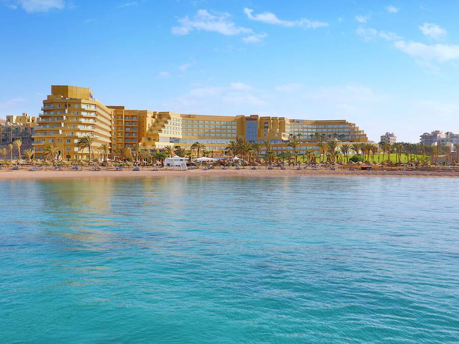 A hotel with multiple floors, a beach in the foreground, and palm trees.