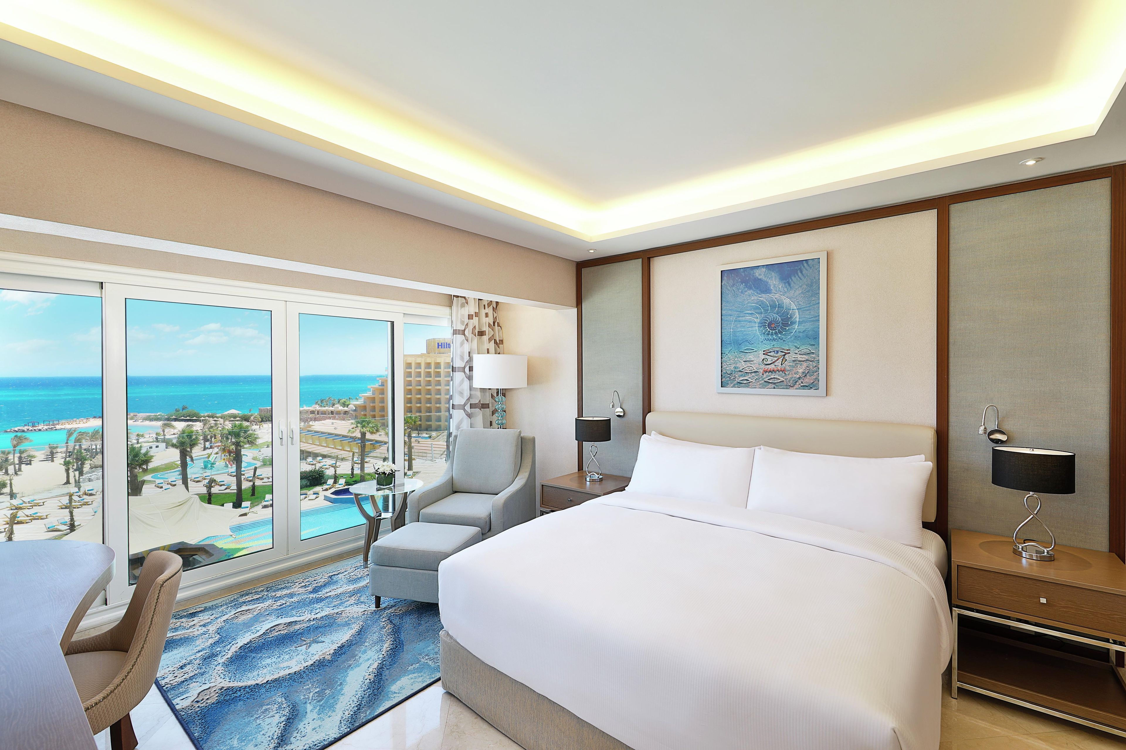 A hotel room with a large bed, a chair, a desk, and a view of a beach and pool.