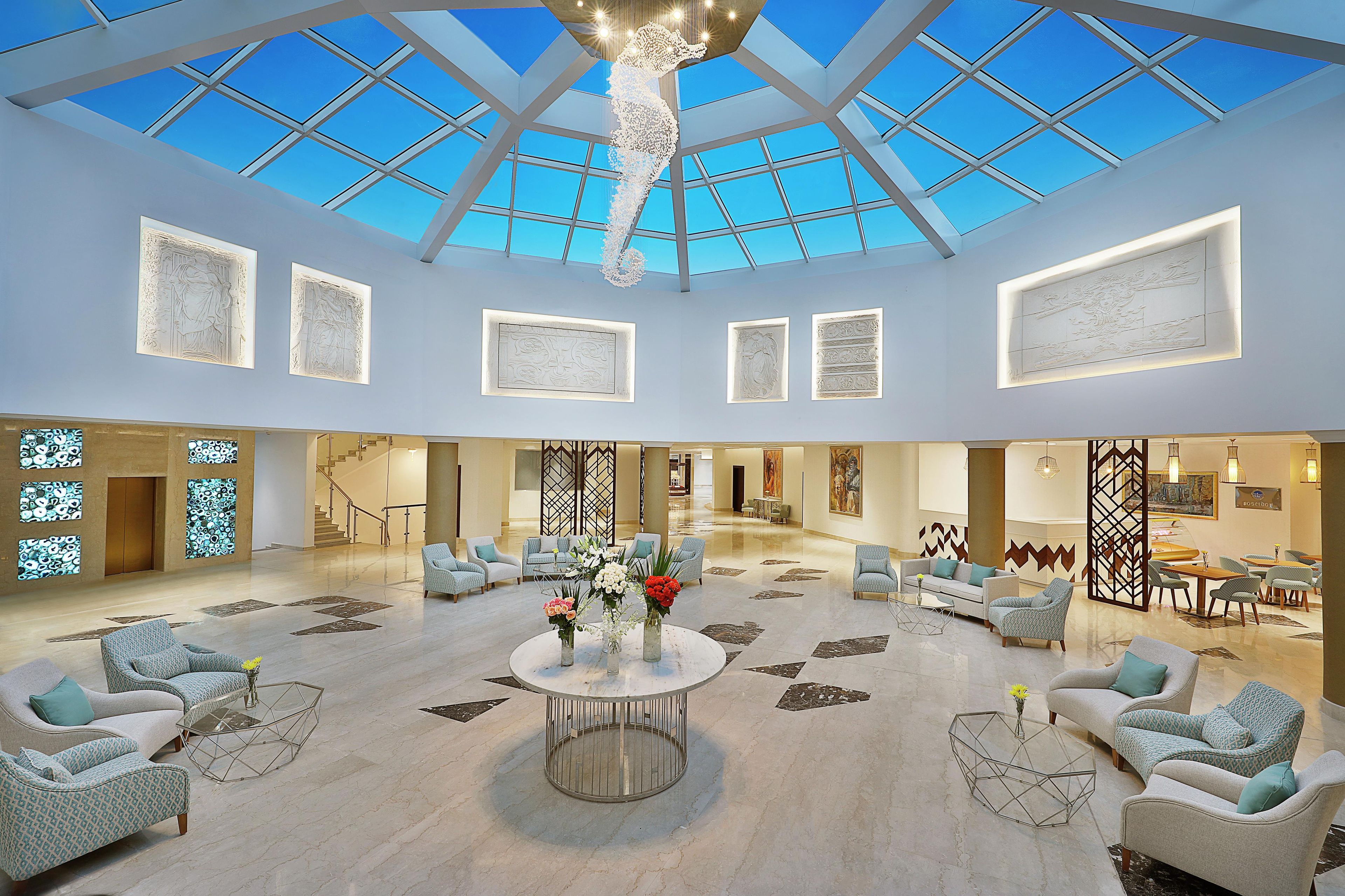 Een ruime lobby met een hoge plafond, grote ramen en een centraal zithoek met ronde tafels en stoelen.