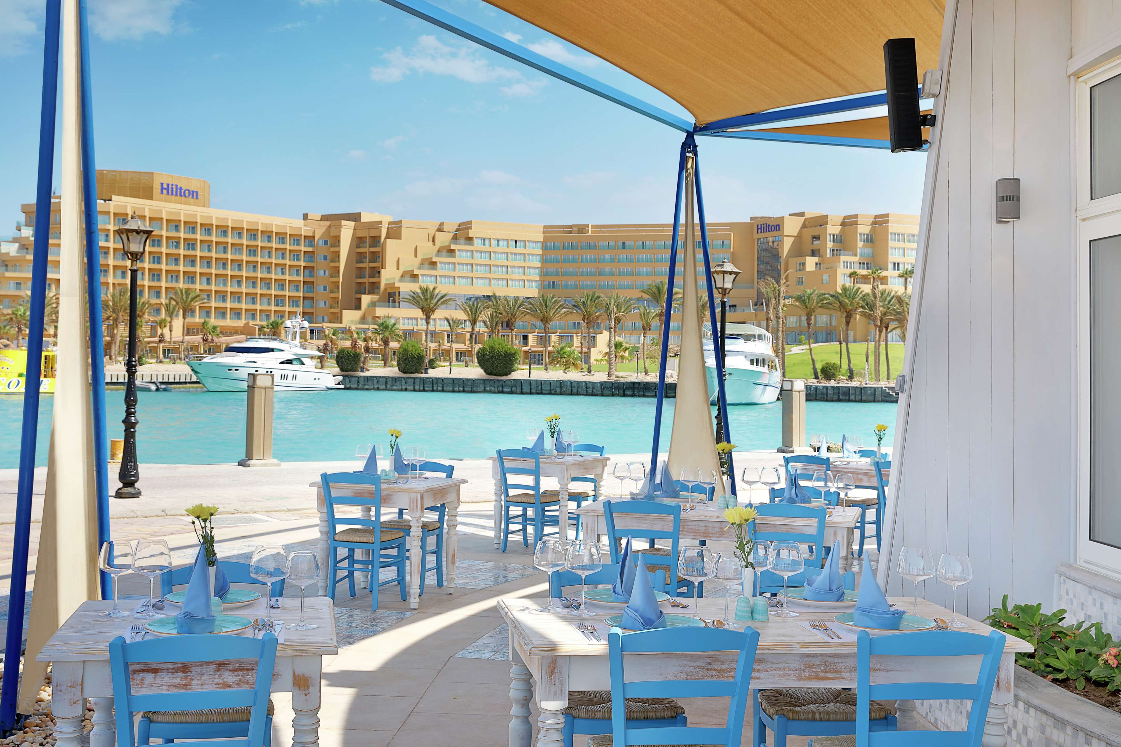 Un área de comedor al aire libre con mesas y sillas, con vistas a un puerto deportivo con barcos y un hotel al fondo.