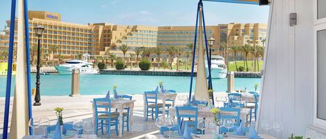 Un área de comedor al aire libre con mesas y sillas, con vistas a un puerto deportivo con barcos y un hotel al fondo.