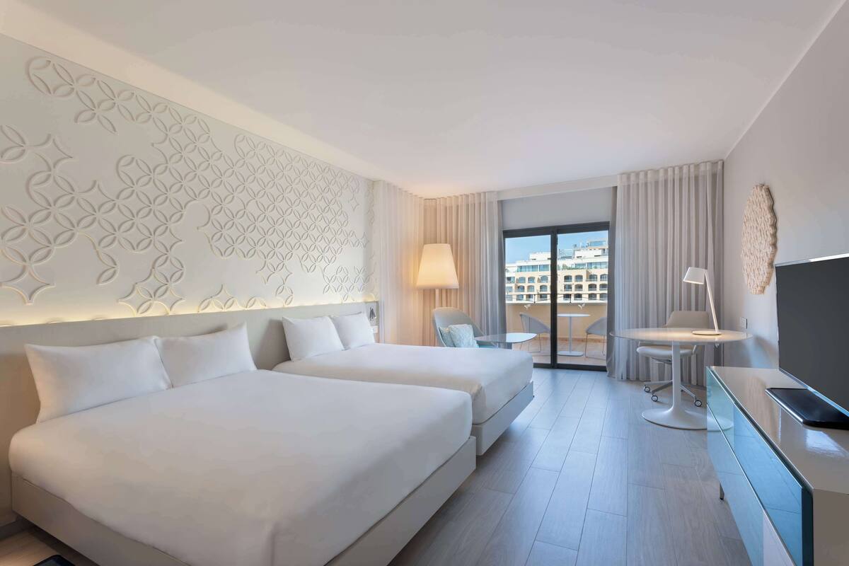 Une chambre d’hôtel avec deux lits, un bureau, une chaise, une télévision et une vue sur les immeubles par la fenêtre.